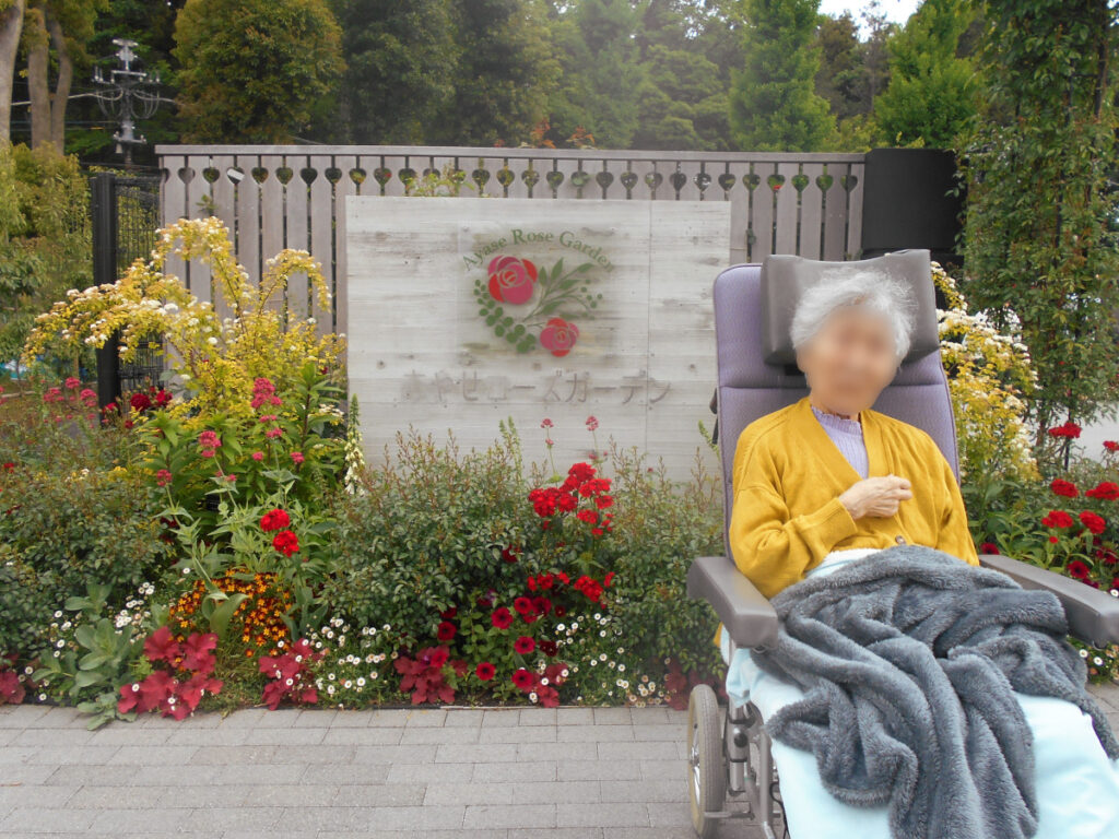 特別養護老人ホーム　ル・リアンふかみの外出レクリエーション。あやせローズガーデンのエントランスの前にいるご利用者様です。エントランス前の看板は、周りにいろいろな種類の花が咲いています。