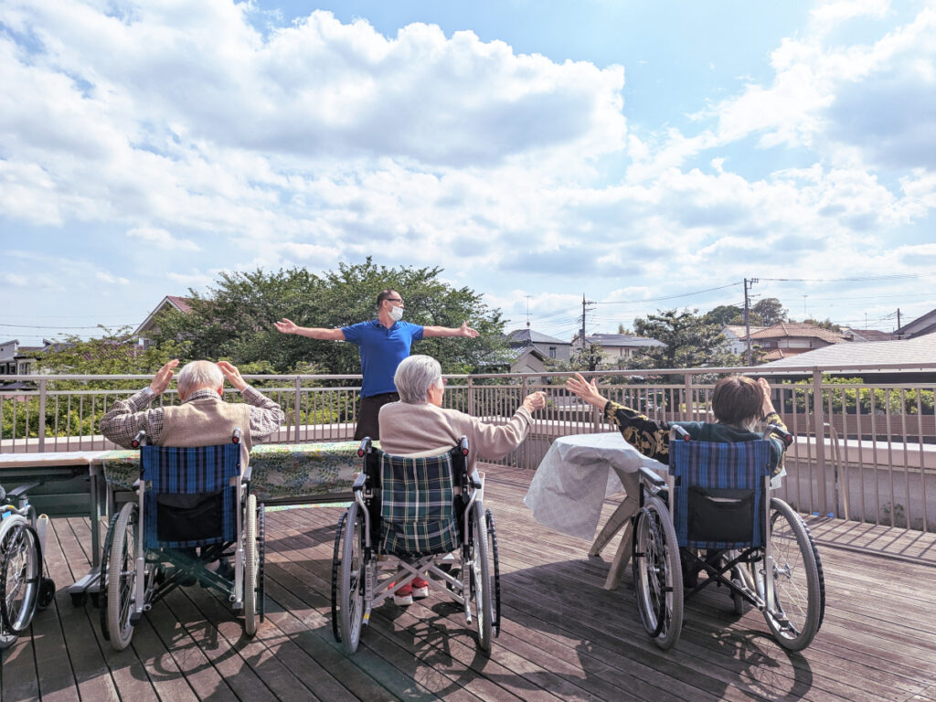 特別養護老人ホーム　ル・リアンふかみ2階のウッドデッキにて、体操をする施設職員とご利用者様。晴天の青空が広がっています。