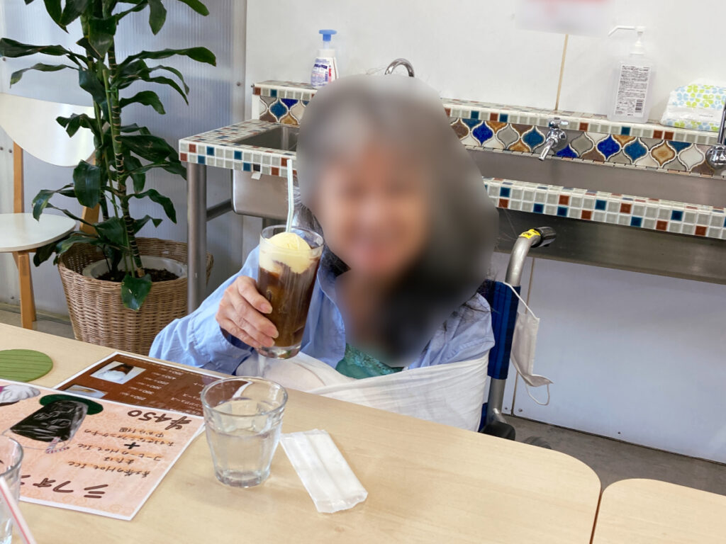 特別養護老人ホーム　ル・リアンふかみの外出レクリエーションの様子です。カフェの席で、コーヒーフロートを持って笑顔のご利用者様です。