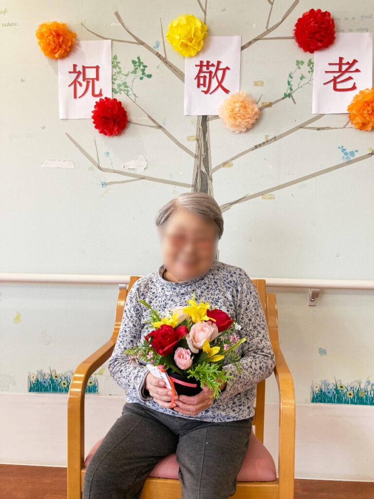 特別養護老人ホーム ル・リアンふかみで、令和8年9月17日(木)に行った『敬老の日』のレクリエーションの様子です。お花を持って、笑顔で記念撮影をするご利用者様です。